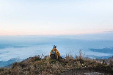 Genç bir gezgin dağın tepesinde oturmuş güzel bir gündoğumunda sisli bir denizle kaplı manzarayı seyrediyor. Dağın tepesindeki maceraperest adamın dikiz görüntüsü.