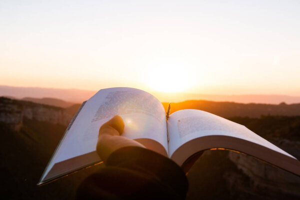 Anonymous person hand holding a book on a beautiful landscape at sunset or sunrise background. Concept of going out into nature to disconnect as a source of inspiration.