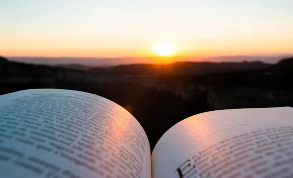 Reading adventure travel book in the mountain with sunset or sunrise in the background. Open book with defocused unreadable text. Concept of stories, imagination, disconnecting from the routine.