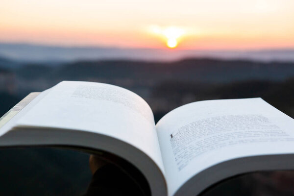 Reading a book in the mountain with sunset or sunrise in the background. Open book with defocused unreadable text. Concept of stories, travel, dreaming, imagination and disconnecting from the routine.