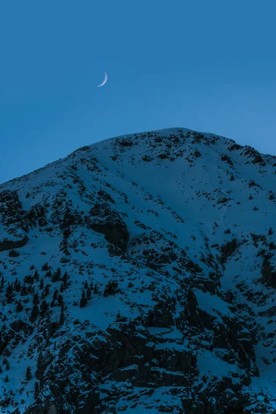 Snowy mountain peaks with the moon at blue hour. Nature background landscape. Moon over the snowy mountains with blue and clear sky on early morning in the mountains. Vertical wallpaper.
