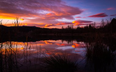 Göl suyuna yansıyan turuncu ve kırmızı bulutlarla muhteşem bir gün batımı. Güneş doğarken doğal ayna. Estany de Banyoles, Girona, Katalonya, İspanya.