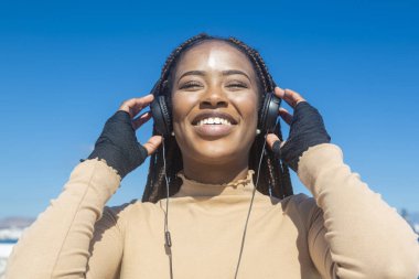 Kulaklık ve mavi gökyüzü geçmişiyle gülümseyen genç siyah afro kızın yakın plan portresi, pozitiflik ve huzur saçan güzel afro kız..