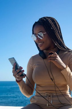 Genç siyah afro kadın cep telefonuna bakarak gülümsüyor, açık havada mavi gökyüzü ve deniz altyapısı ile. Teknoloji, iletişim, sosyal ağlar.