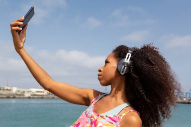 afro woman with headphones taking a selfie with mobile phone in the background seascape