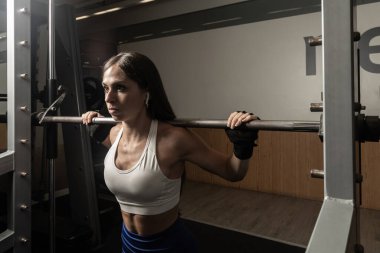 Atletik kadın bir demirci makinesinde çömelme hareketleri yapıyor, spor salonuna konsantre oluyor.