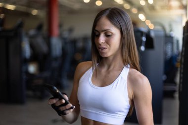 Cep telefonu kullanan atletik bir kadın spor salonunda egzersize ara veriyor.
