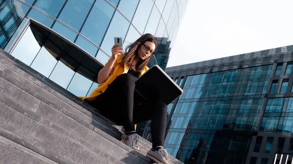 Parlak sarı ceketli genç bir kadın dizüstü bilgisayar ve akıllı telefon kullanıyor..