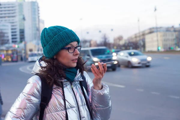 Senden Sie eine Sprachnachricht im Messenger. Eine junge Frau in Daunenjacke und Hut telefoniert, — Stockfoto