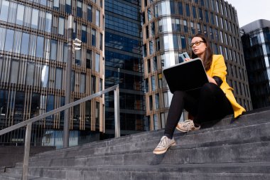 Parlak sarı ceketli genç bir kadın dizüstü bilgisayar ve akıllı telefon kullanıyor..