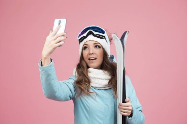 Eğlenceli kayakçı akıllı telefonuyla selfie çekiyor. Ceketli ve şapkalı genç bir kadın.,