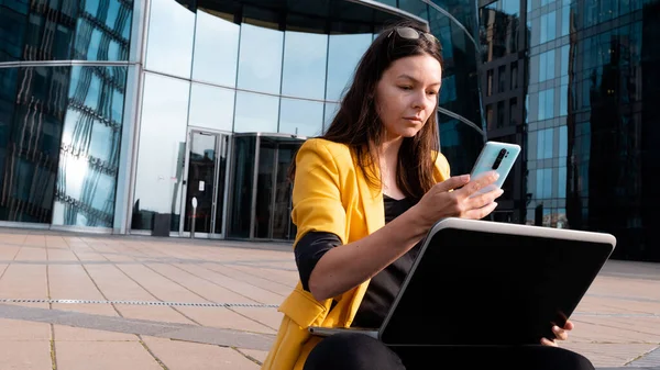Fiatal nő egy fényes sárga kabát használ laptop és okostelefon. — Stock Fotó