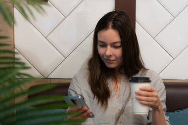 Genç bir kadın bir kafede oturuyor, karton bardakta kahve içiyor.