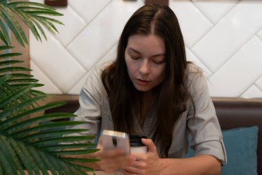 Genç bir kadın bir kafede masada oturuyor ve mobil uygulamalarla internetten iletişim kuruyor.,