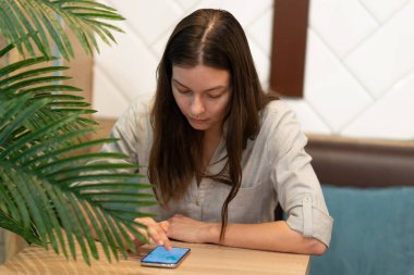 Genç bir kadın bir kafede masada oturuyor ve mobil uygulamalarla internetten iletişim kuruyor.,