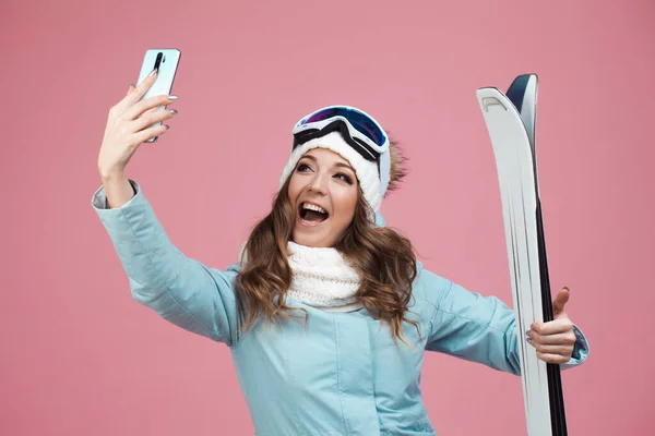 Skieuse amusante prend un selfie sur son smartphone. Une jeune femme avec du matériel de ski en veste et chapeau, — Photo