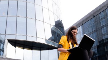 Parlak sarı ceketli genç bir kadın dizüstü bilgisayar ve akıllı telefon kullanıyor..