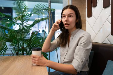 Genç bir kadın bir kafede masada oturuyor ve mobil uygulamalarla internetten iletişim kuruyor.,