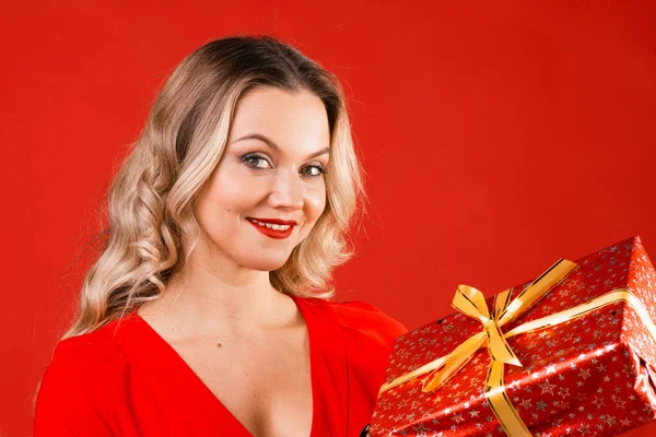 Give gifts for the holiday. Cheerful young woman in a red dress with a red gift box in her hands.