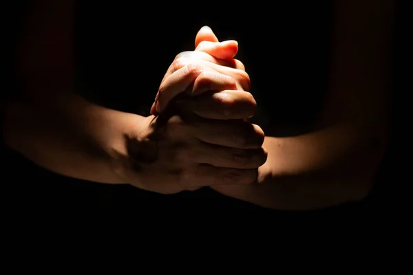 Rezando mãos cruzadas sobre um fundo preto. Luz de cima. Mãos dobradas em oração. — Fotografia de Stock