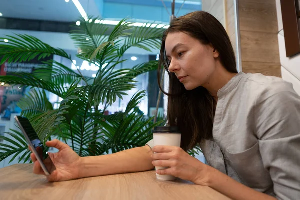 Genç bir kadın bir kafede masada oturuyor ve mobil uygulamalarla internetten iletişim kuruyor.,