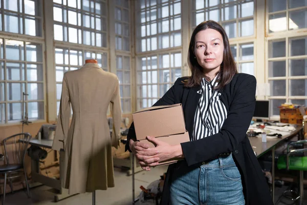 Kleinbedrijf, kleermaker winkel, kostuum ontwerpers workshop. Een jonge vrouwelijke ondernemer houdt meerdere dozen in haar handen. — Stockfoto