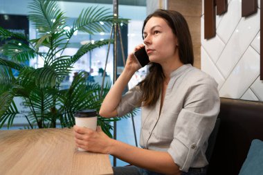 Genç bir kadın bir kafede masada oturuyor ve mobil uygulamalarla internetten iletişim kuruyor.,