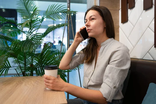 Genç bir kadın bir kafede masada oturuyor ve mobil uygulamalarla internetten iletişim kuruyor.,