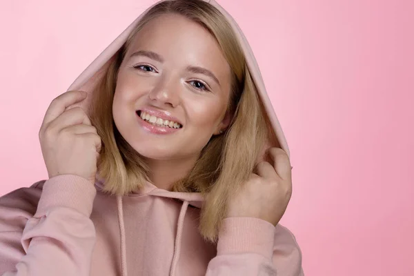 Une jeune blonde mignonne avec une capuche sur la tête, dans un sweat à capuche rose sur un fond rose — Photo