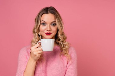 Çay iç, sıcak bir içeceğin tadını çıkar. Pembe arka planda, kalın pembe kazaklı genç bir kadın.