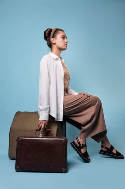 Yazlık pantolonlu ve beyaz gömlekli mutlu genç bir kadın klasik deri bavulların üzerinde oturuyor.
