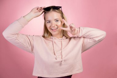 Alnında güneş gözlüğü olan çekici genç bir sarışın zafer işareti gösteriyor.,