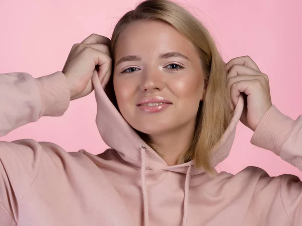 Mladá roztomilá blondýna s kapucí na hlavě, v růžové mikině na růžovém pozadí — Stock fotografie