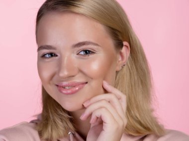 Pembe kapüşonlu tatlı sarışın mutlu bir şekilde gülümseyip eliyle yüzüne dokunuyor..