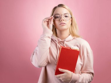 Pembe kapüşonlu ve gözlüklü genç bir öğrenci elinde bir kitap tutuyor ve bir konu üzerinde çalışıyor.,