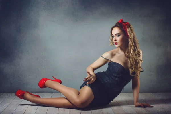 Sexy blonde vrouw in jeans sundress en rode schoenen pin up meisje retro vrouw sexy benen en een rode capitium op zijn hoofd op de vloer zittend op de vloer kruipen en rechtzetten haar — Stockfoto