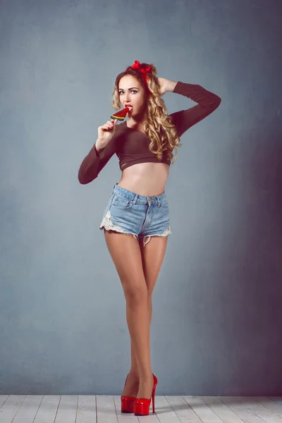 Beautiful young sexy blonde slim figure with a red armband with red lips in denim shorts in red high heels with long legs of sensual temptation watermelon candy emotion surprise delight Pin Up — Stock Photo, Image