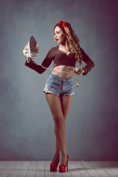 Pin up Young beautiful sexy girl with make-up blonde with a red band in denim shorts housewife with iron in red shoes slender legs — Stock Photo, Image