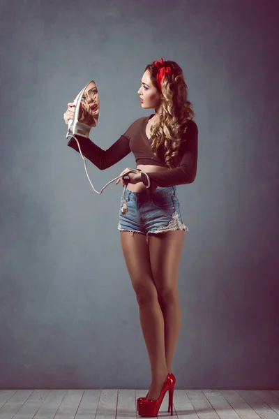 Pin up Young beautiful sexy girl with make-up blonde with a red band in denim shorts housewife with iron in red shoes slender legs — Stock Photo, Image