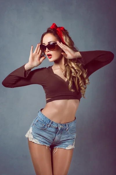 Hermosa joven sexy rubia figura delgada con un brazalete rojo con labios rojos en pantalones cortos de mezclilla en tacones altos rojos con piernas largas con gafas Pin Up culo —  Fotos de Stock