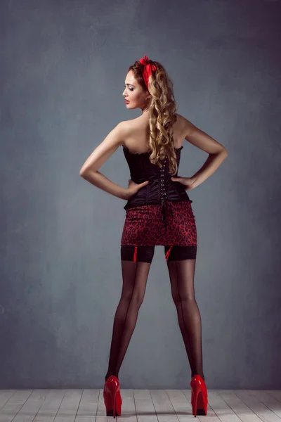 Pin up sexy mooie jonge blonde vrouw make-up permanent in een korset in een korset en mini rok, zwarte kousen op hoge hakken rode schoenen — Stockfoto