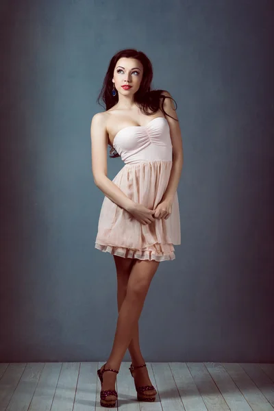 Sexy young beautiful brunette with long hair, with a slender figure standing on a lifting leg up holding his heel in a pink dress and retro women athletic and acrobatic pin up make-up — Stock Photo, Image