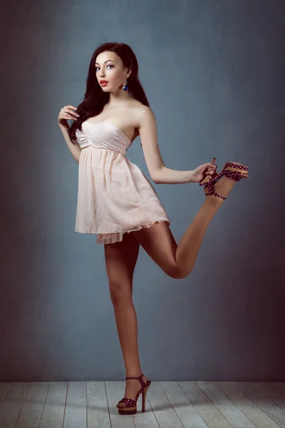Sexy young beautiful brunette with long hair, with a slender figure standing on a lifting leg up holding his heel in a pink dress andretro woman athletic and acrobatic pin up  make-up — Stock Photo, Image