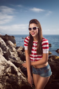 Genç güzel kız kısa kot pantolon ve uzun saç kayalar gözlük martılar su bulutlar, dalga, rüya artistik gözlüklü olan çizgili tişört denizde üzerinde duygular pin, diş ile açık ağız gülümse