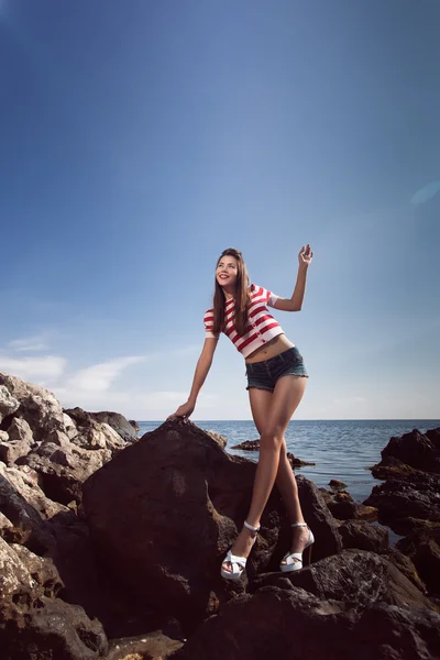 Épingler jeune belle fille sur la mer en jeans courts et T-shirt rayé avec des cheveux longs rochers lunettes goélands nuages d'eau, vague, figure de rêve avec des lunettes émotions, sourire avec des dents, bouche ouverte — Photo