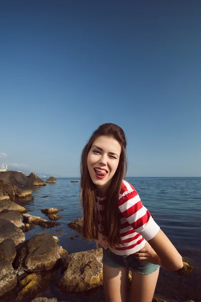 Épingler jeune belle fille sur la mer en jeans courts et T-shirt rayé avec des cheveux longs rochers lunettes goélands nuages d'eau, vague, figure de rêve avec des lunettes émotions, sourire avec des dents, bouche ouverte — Photo