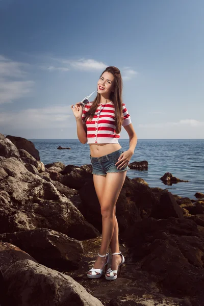 Pin up young beautiful girl on the sea in short jeans and striped T-shirt with long hair rocks glasses gulls water clouds, wave, dream figure with glasses emotions, smile with teeth, open mouth — Stock Photo, Image