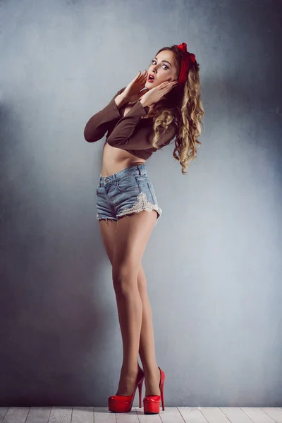 Bonita jovem loira sexy figura slim com uma braçadeira vermelha com lábios vermelhos em calções jeans em saltos altos vermelhos com pernas longas Pin Up — Fotografia de Stock