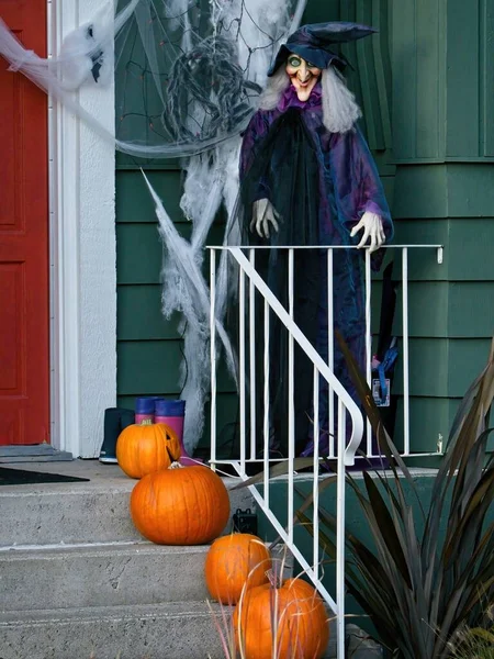 Full-size witch doll stands at the porch of the house as a halloween decoration. Pumpkins decorate the steps