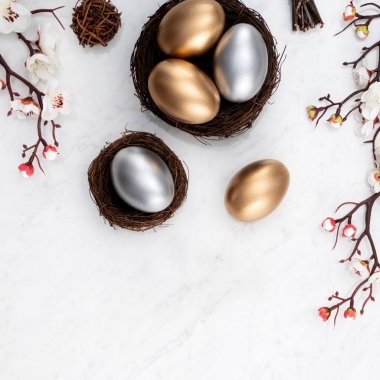 Yuvasında altın ve gümüş Paskalya yumurtaları, parlak mermer masa arkasında beyaz erik çiçekleri olan tasarım konsepti..
